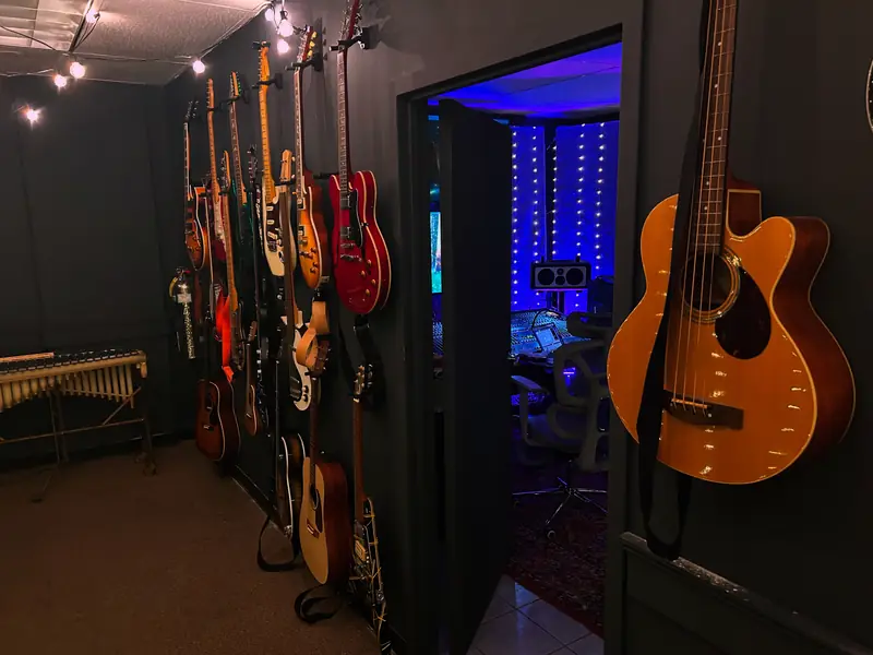 Wall of electric and acoustic guitars and basses, with the control room visible through the doorway